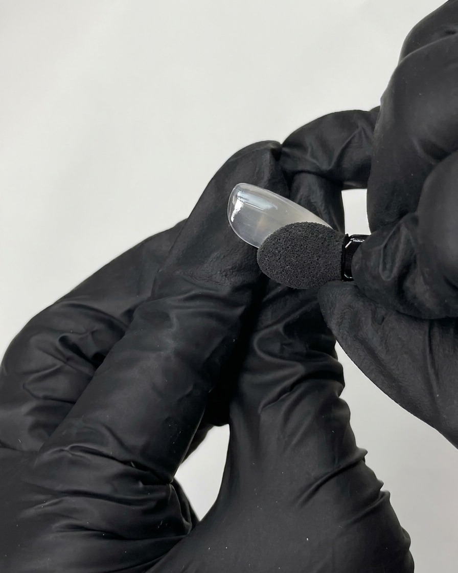 Una mano con guante negro usando un aplicador de esponja para frotar polvo cromado iridiscente sobre la superficie de una punta de uña postiza lisa.