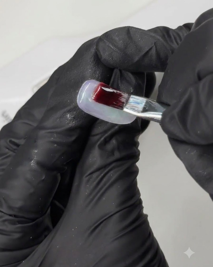 Un pincel aplicando un gel profundamente pigmentado sobre una punta de uña de práctica durante un tutorial de arte de uñas de múltiples pasos.