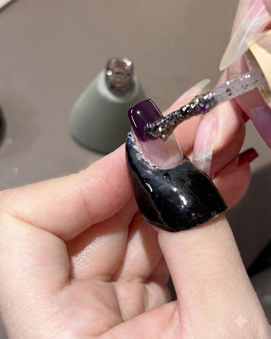 Un pincel aplicando gel con purpurina morada sobre una punta de uña morada oscura, tirando de él ligeramente hacia arriba.