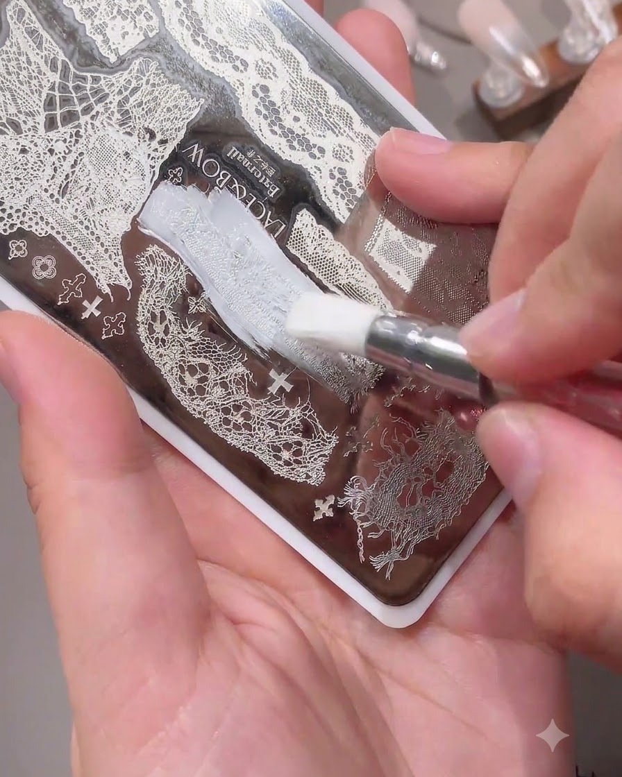 Pincel aplicando esmalte blanco sobre un delicado patrón de encaje en una placa de metal para estampación de uñas.