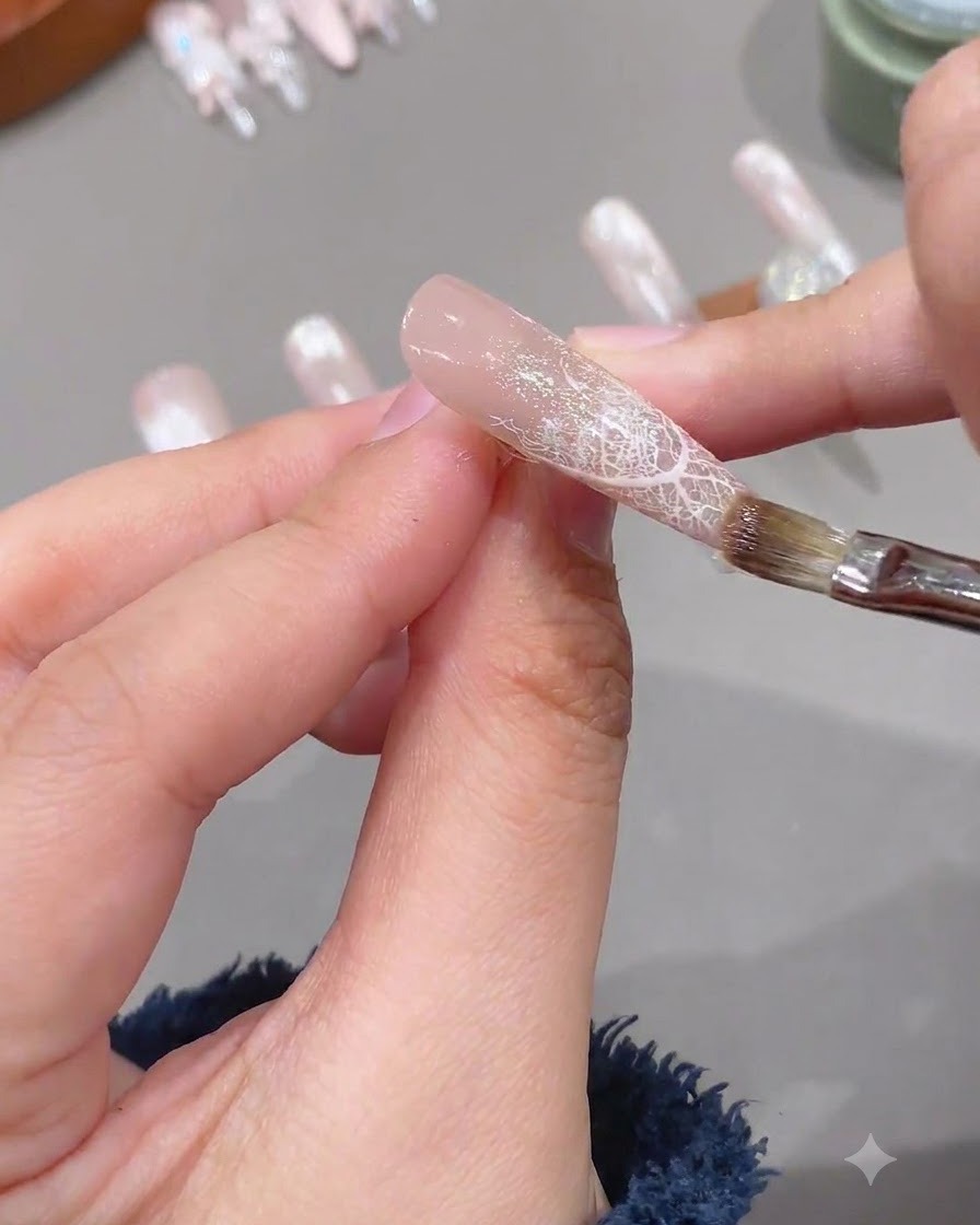 Pincel aplicando una capa transparente de gel de refuerzo suave sobre una uña preparada con diseño de encaje.