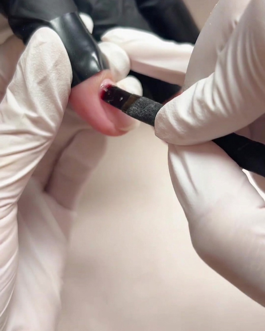 Una vista de cerca de una mano con guantes usando un pincel pequeño para aplicar base de gel transparente sobre una uña natural.
