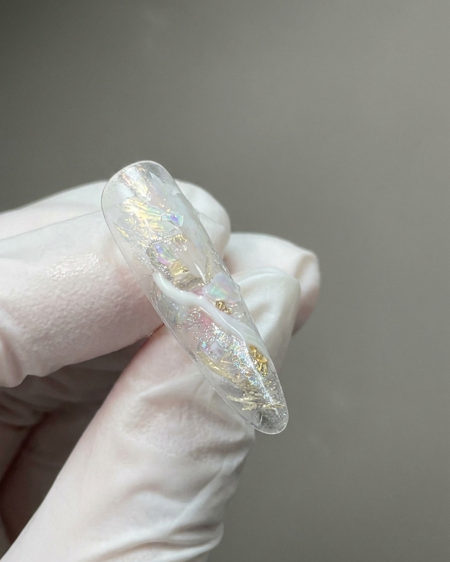 Un primer plano de una punta de uña transparente que presenta escamas brillantes incrustadas y mechones de detalles de mármol blanco lechoso.