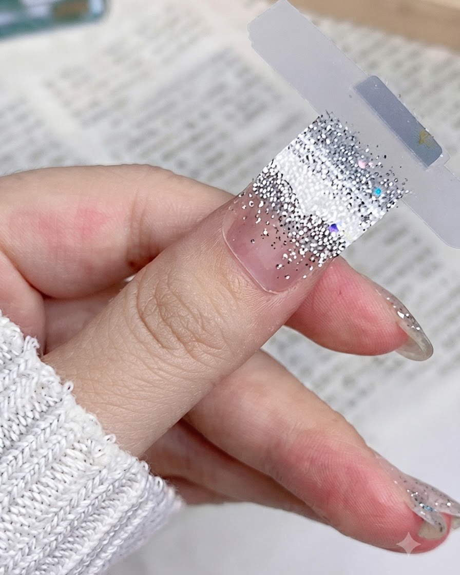 Un primer plano de una pegatina de uñas con purpurina plateada colocada de forma plana y suave sobre la uña natural del pulgar.