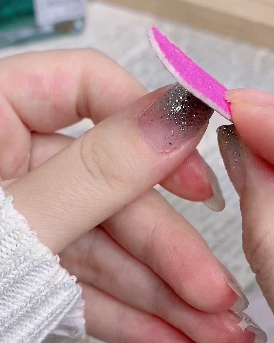 Una lima de uñas rosa limando la parte sobrante de una pegatina de purpurina plateada en un pulgar.
