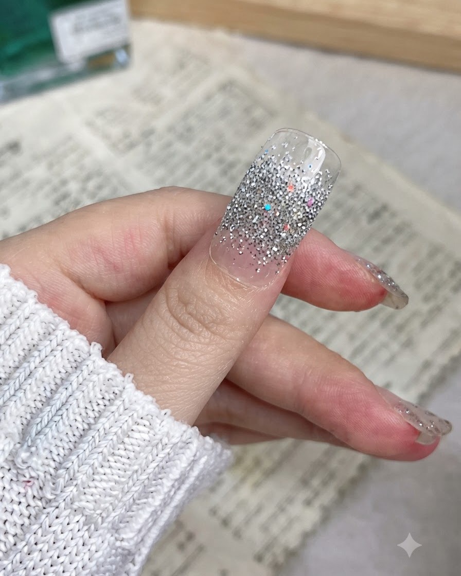 Un primer plano de un pulgar con una pegatina de uñas con brillo plateado aplicada suavemente desde la cutícula hasta el borde.
