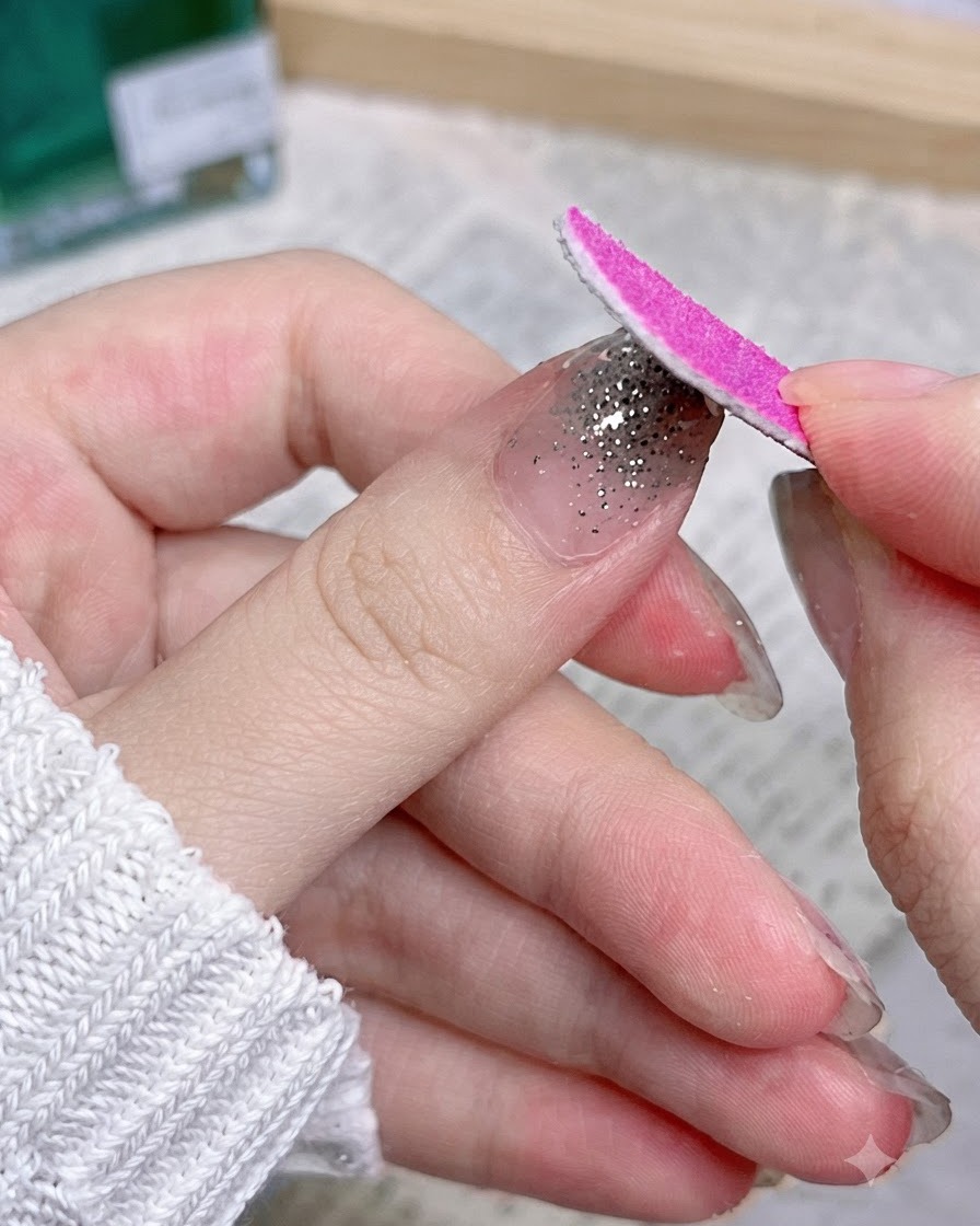 Una lima de uñas rosa moviéndose hacia abajo a lo largo del borde de una uña para eliminar el exceso de material de la pegatina.