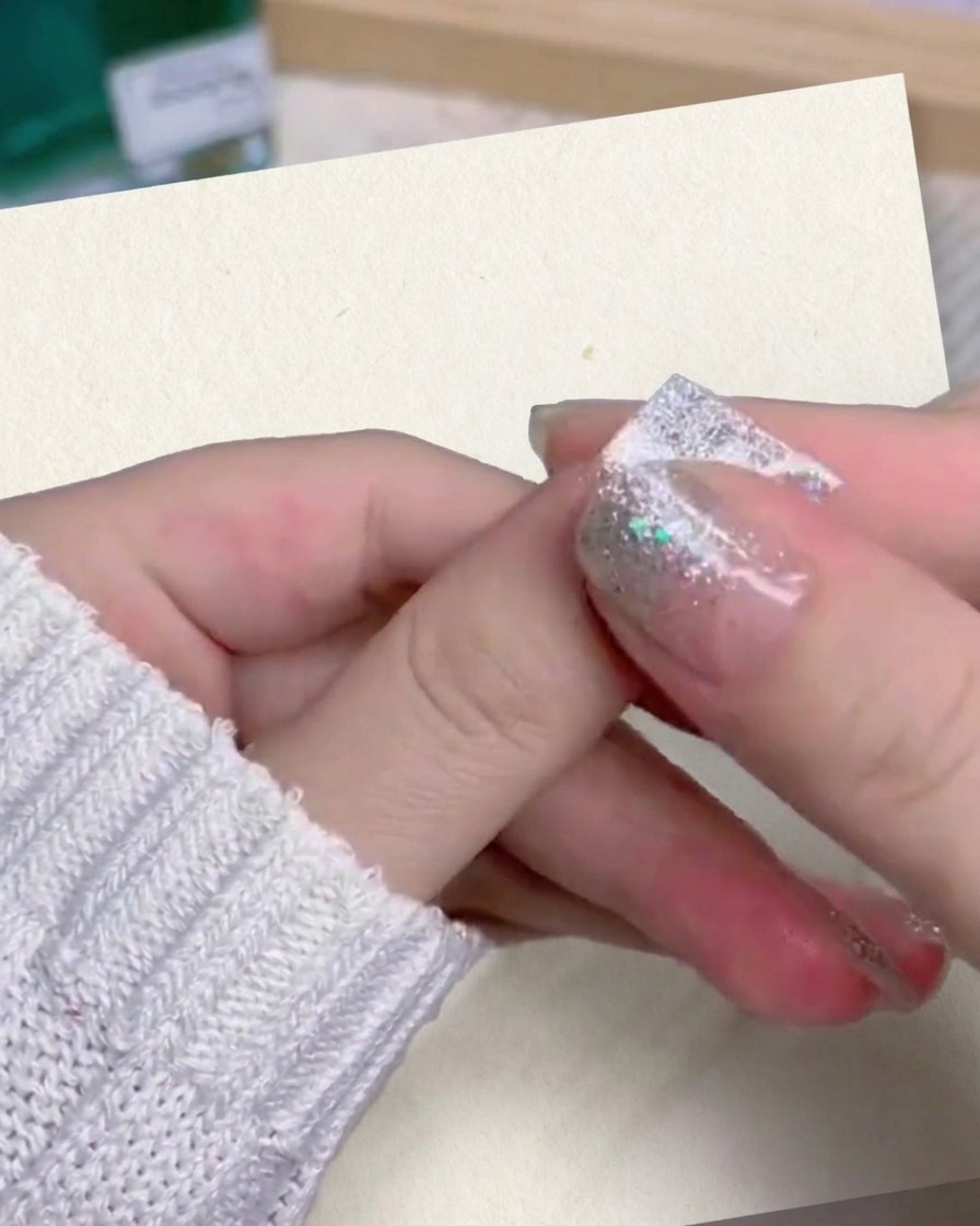 Un primer plano del pulgar y el dedo índice de una persona presionando y alisando firmemente una uña postiza con degradado de purpurina plateada en su dedo.