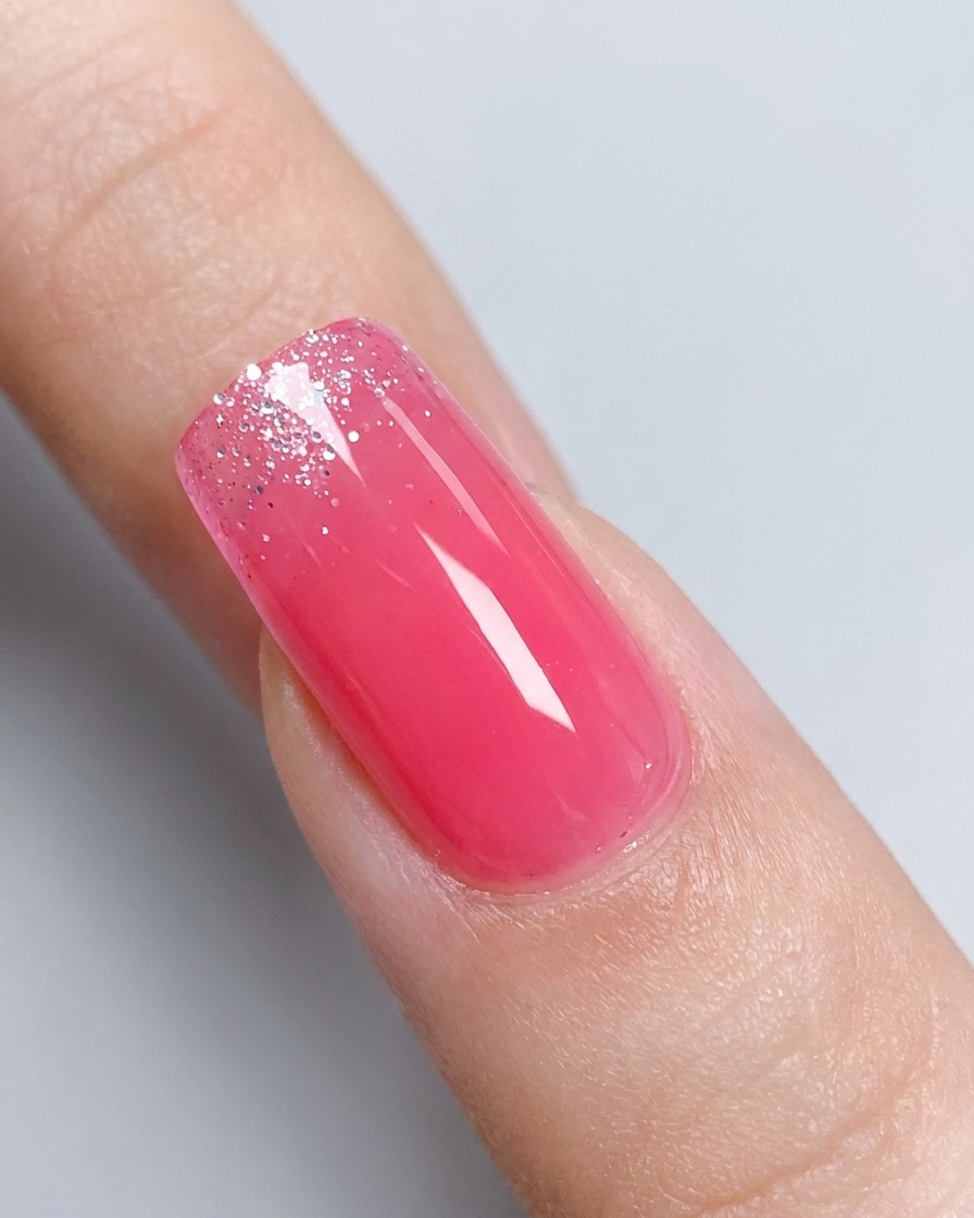 Un primer plano de una uña con degradado rosa brillante y un frasco de gel con glitter plateado ligeramente desenfocado al fondo.