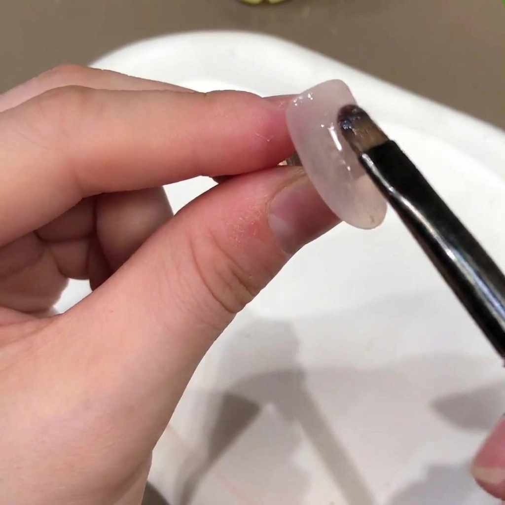 Un pincel aplicando una base de gel de color rosa nude sobre la superficie de una uña postiza.