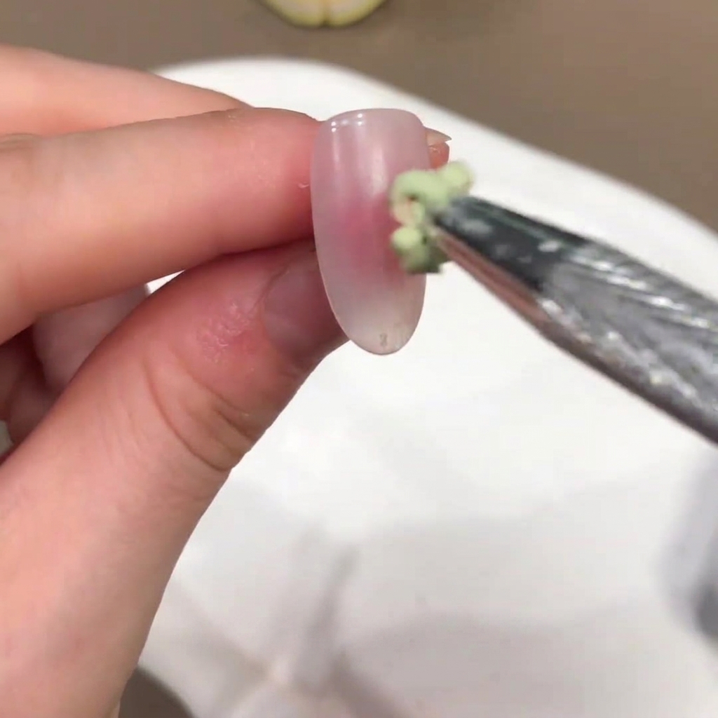 Un pincel pequeño aplicando una gota de gel con un ligero tono verde en el centro de una uña rosa nude.