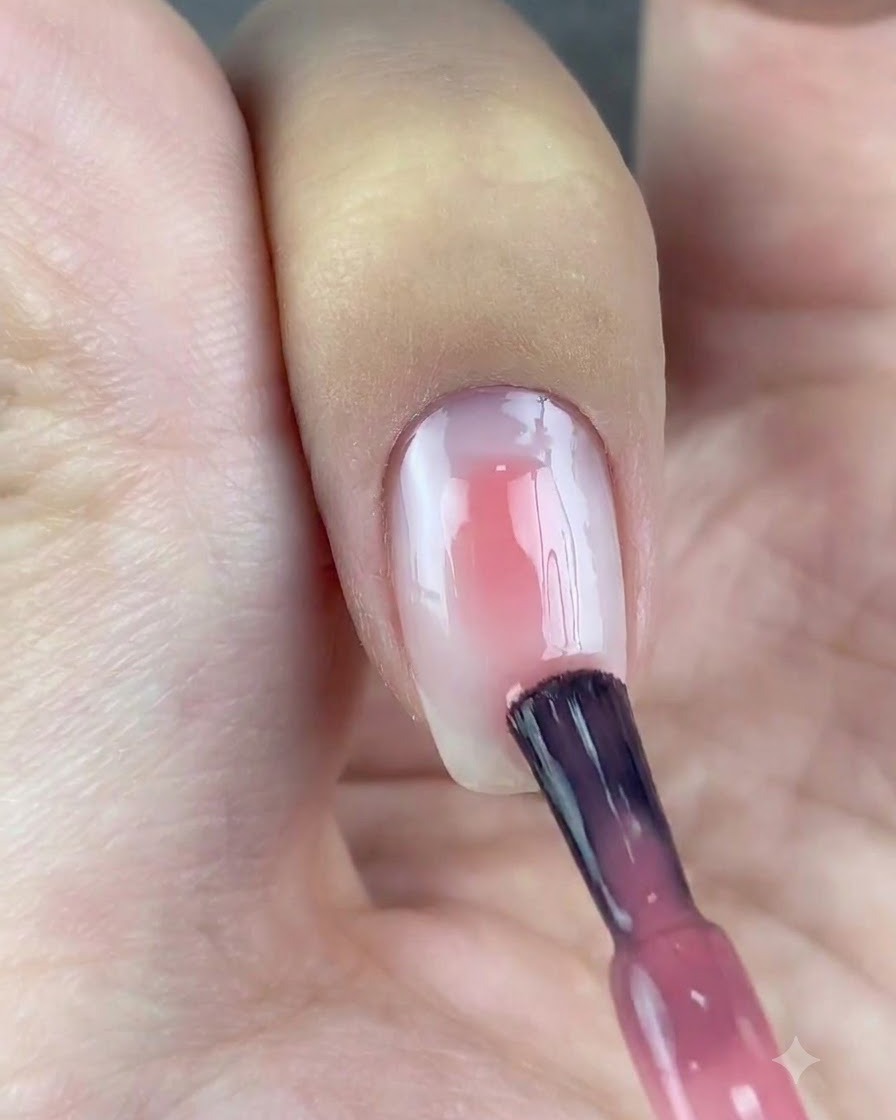 Un pincel de esmalte aplicando una gota de gel rosa translúcido en el centro de una uña con base blanca.