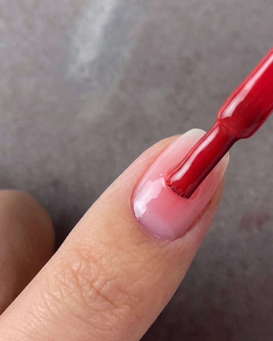 Un pincel de esmalte rojo aplicando una segunda capa de gel rosa en el centro de una uña arreglada.