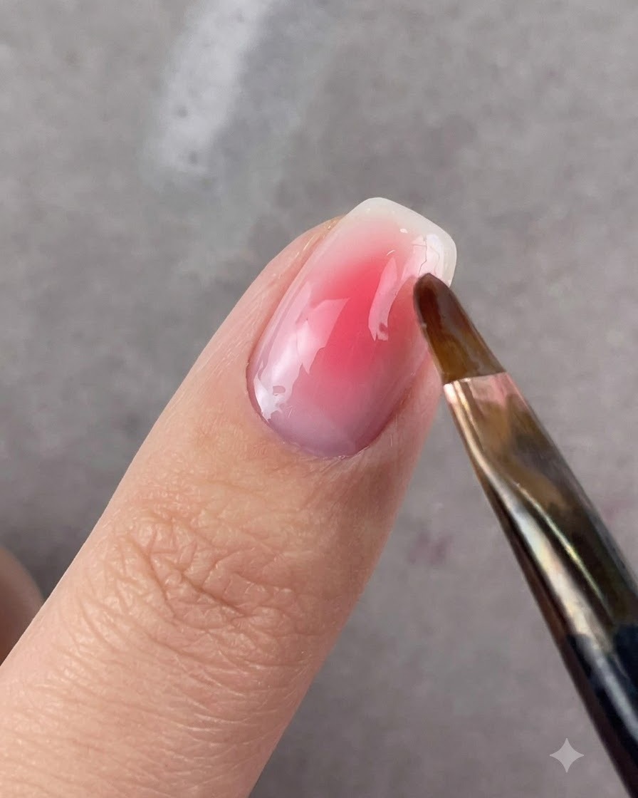 Un pincel de punta redonda difuminando suavemente los bordes del gel rosa en una uña.