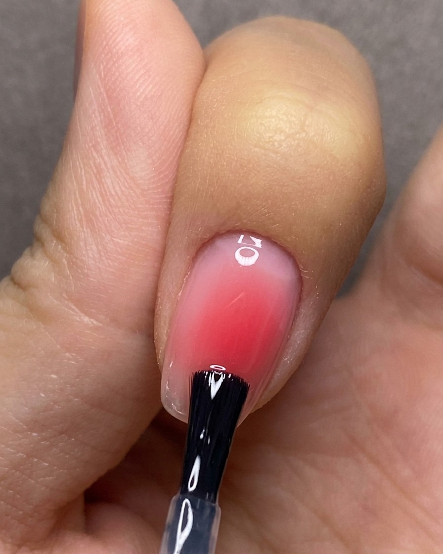 Un pincel aplicando un esmalte final transparente de manera uniforme sobre una uña con diseño degradado rosa.