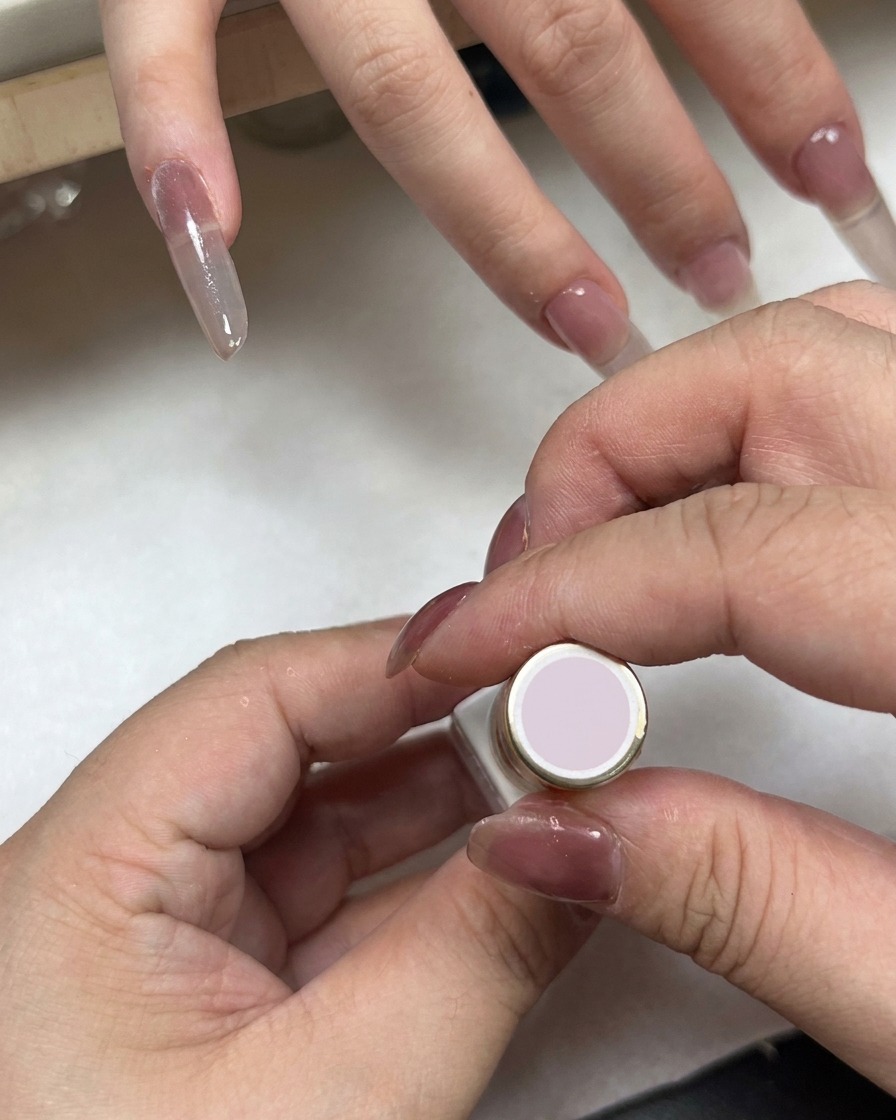 Un frasco de esmalte de uñas con tapa rosa claro sostenido cerca de una mano con uñas en degradado.
