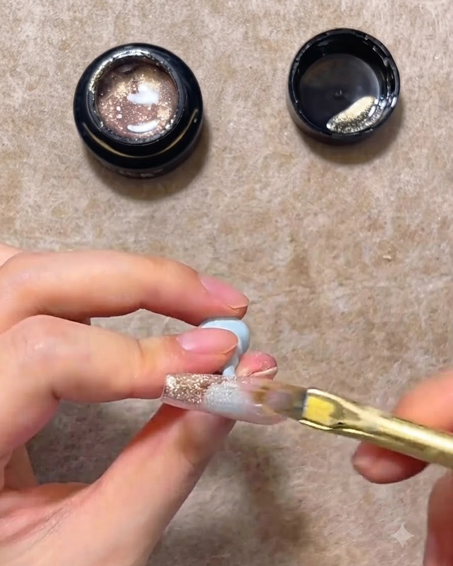 Un pincel de uñas plano barriendo el gel cat eye rosa dorado hacia abajo en una uña postiza transparente para crear un degradado.