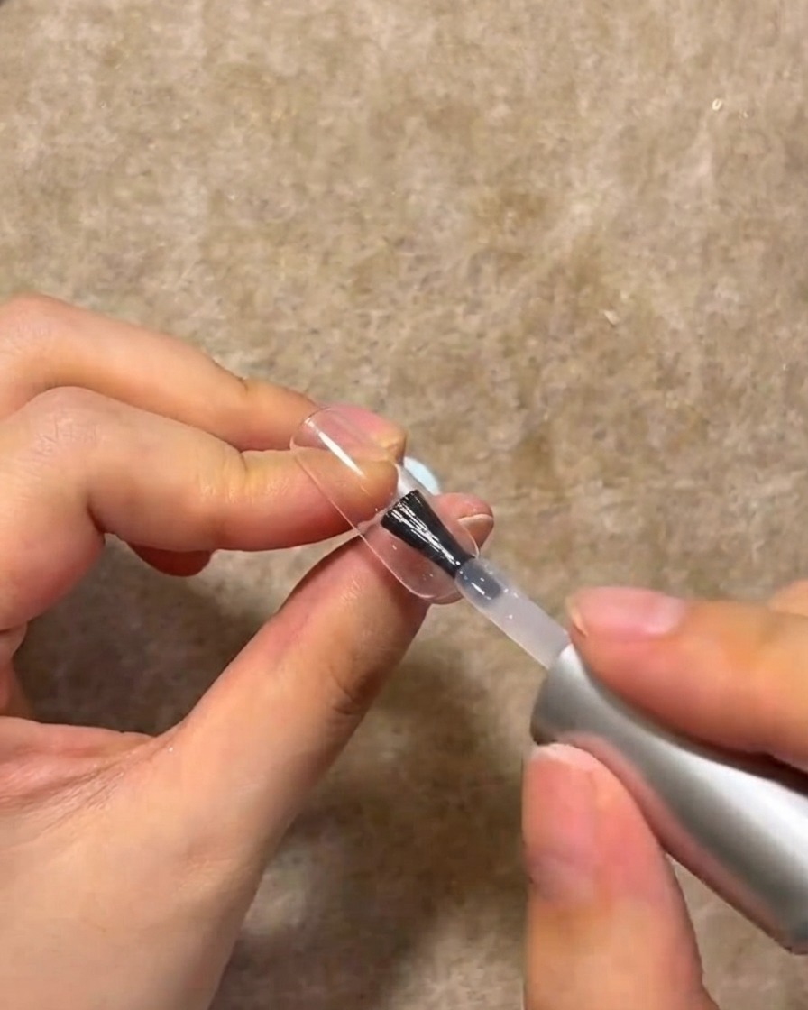 Un pincel de uñas aplicando una capa de gel de construcción transparente sobre la superficie de una uña manicurada.