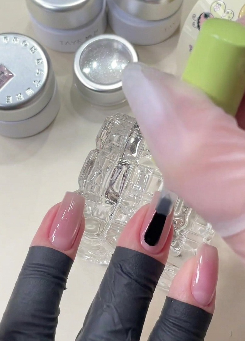 Un pincel aplicando gel base transparente en una uña antes de colocar lentejuelas decorativas en forma de estrella.