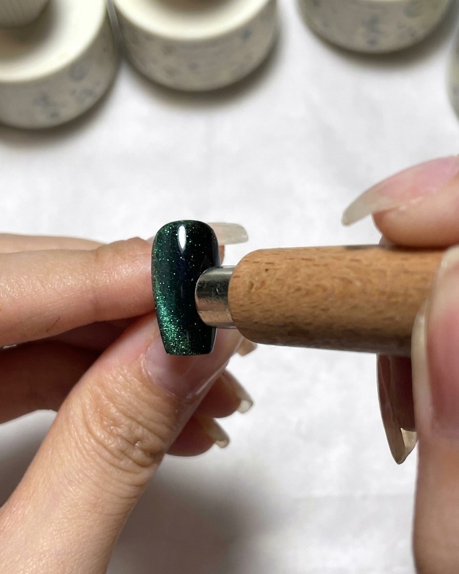 Una herramienta magnética posicionada junto a una uña verde oscuro efecto ojo de gato para distribuir el pigmento.