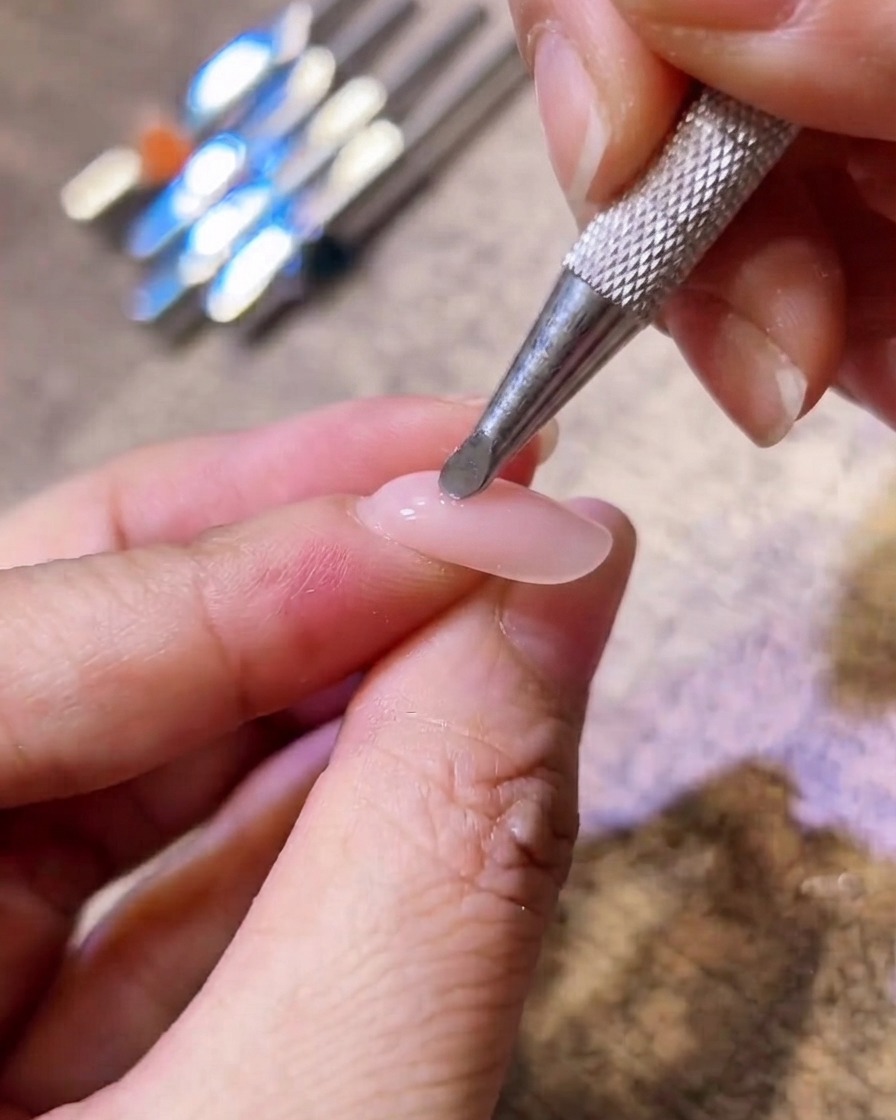 Primer plano de una herramienta metálica aplicando pigmento rosa en el centro de una uña de gel nude.