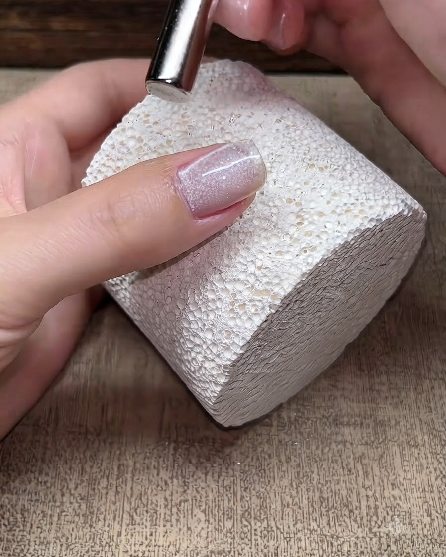 Una pequeña herramienta magnética flotando sobre una uña de gel cat-eye húmeda para manipular las partículas de brillo magnético.