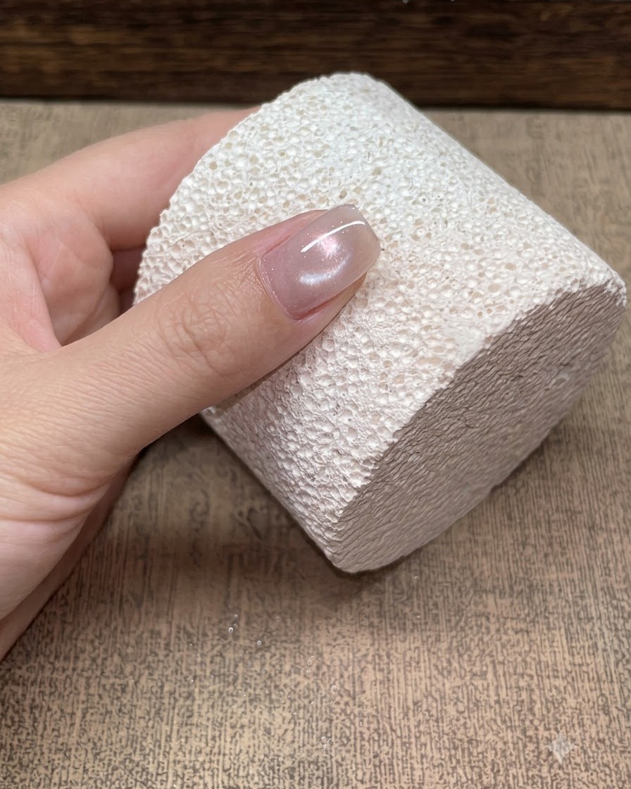 Primer plano de una sola uña apoyada en un bloque blanco texturizado, mostrando un reflejo suave de cat-eye tipo cuenta de vidrio champagne.
