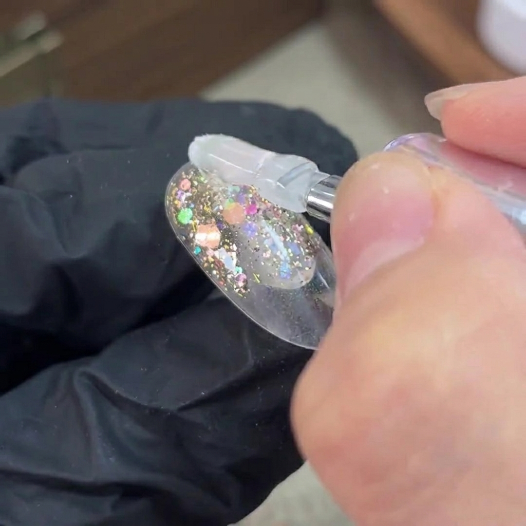 Un pincel aplica gel base transparente sobre una capa de purpurina curada en una uña.