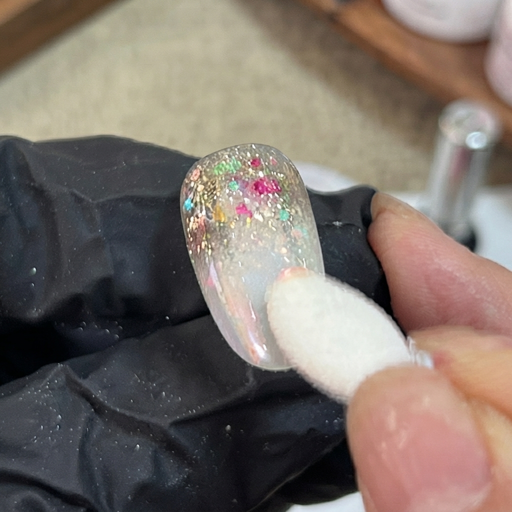 Un aplicador de esponja aplica polvo aurora iridiscente con golpecitos en la sección degradada de una uña artificial decorada.