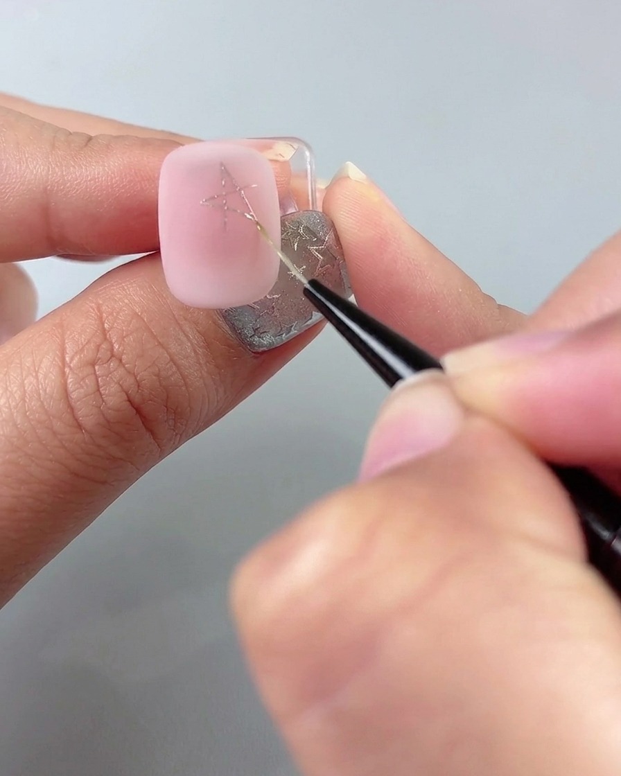 Un pincel de detalles finos dibujando líneas para completar una forma de estrella en una uña rosa manicurada.