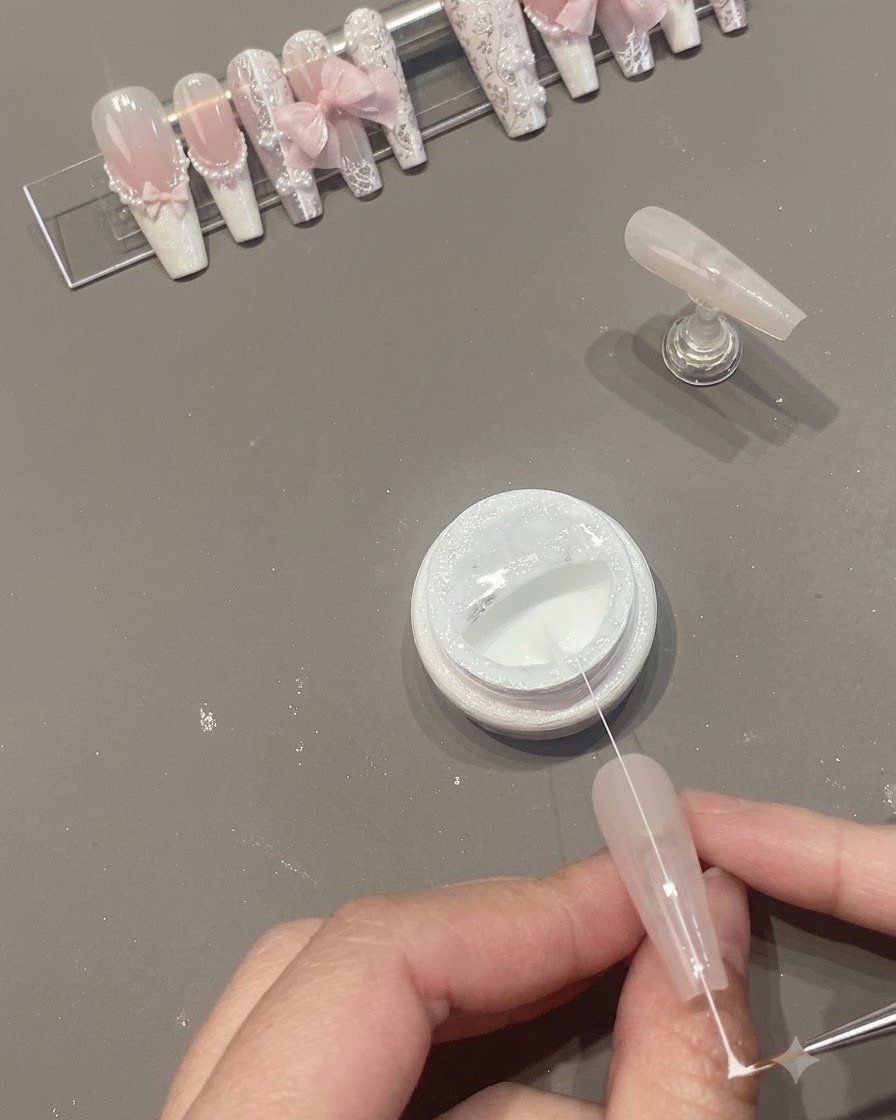 Una hebra estrecha de gel de araña blanco siendo estirada a través de la superficie de la uña sobre un frasco abierto.
