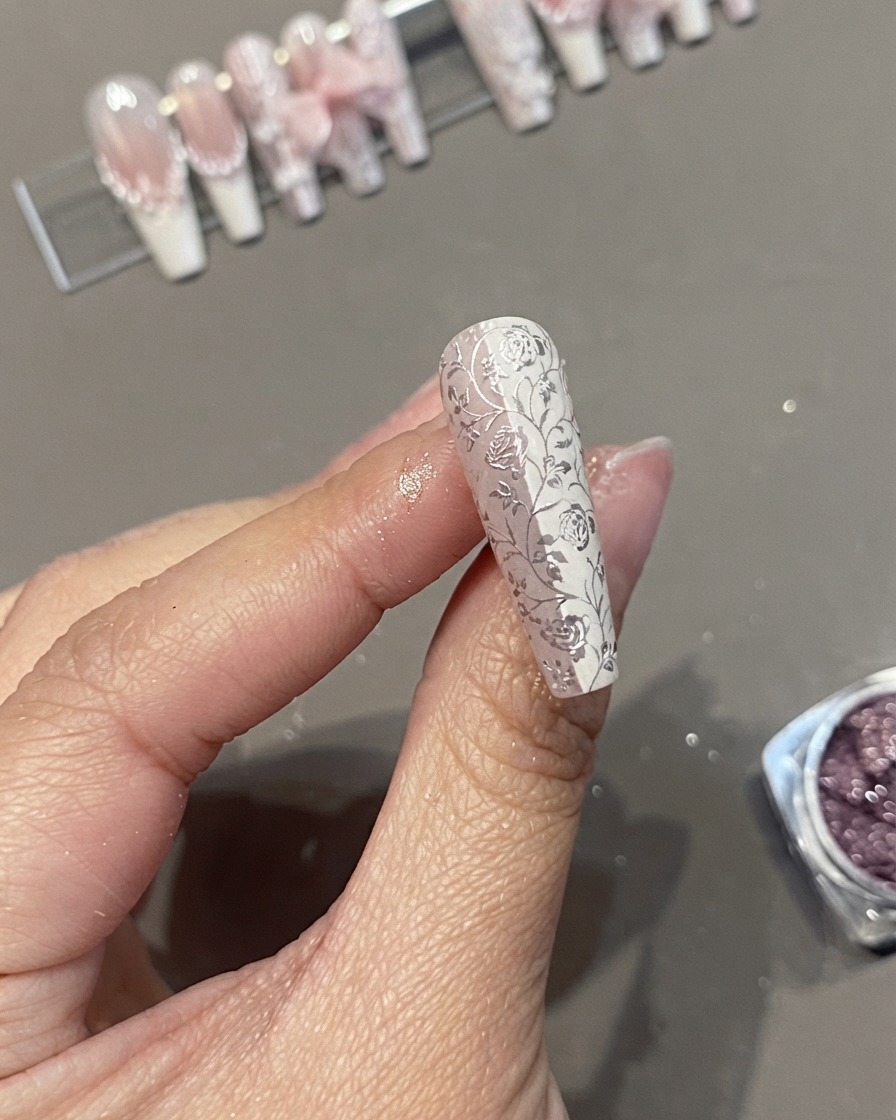 Un primer plano de una uña postiza tipo ataúd larga con un detallado diseño de encaje floral blanco y cromo sostenida entre los dedos.