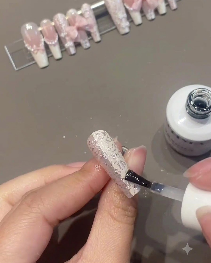 Un pincel de uñas aplicando una capa transparente o superior sobre una uña floral intrincadamente decorada mientras otras uñas terminadas se exhiben en un soporte al fondo.