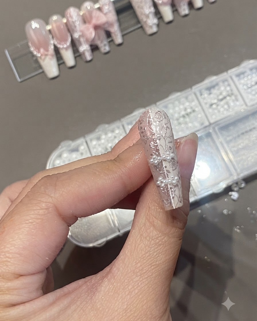 Un pincel de uñas aplicando una capa superior transparente sobre un diseño de uña texturizado que presenta perlas y estampado de encaje.