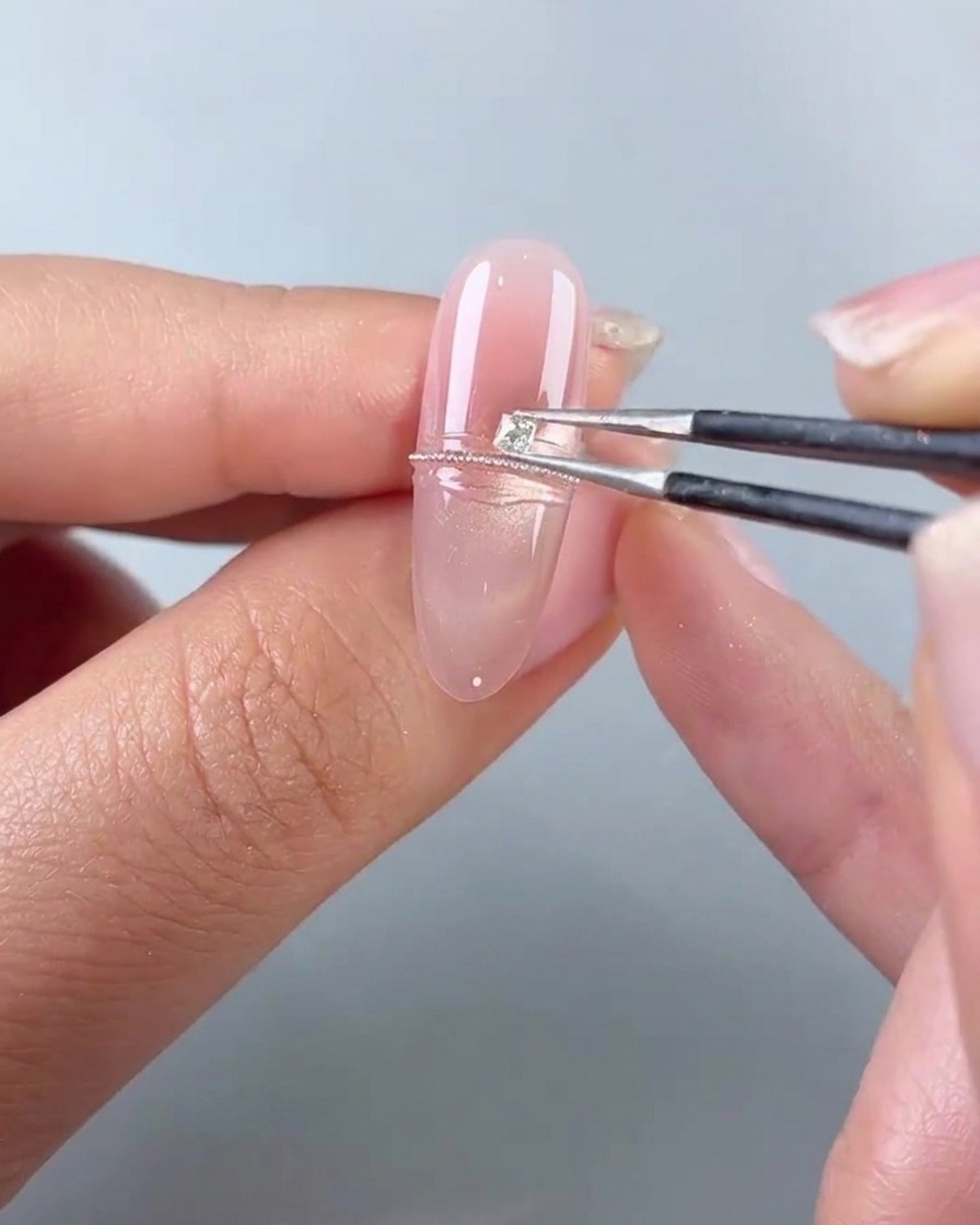 Unas pinzas de precisión colocando un pequeño diamante cuadrado junto a una línea de cuentas de acero en una uña de gel rosa.