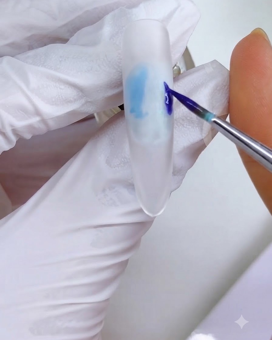 Un pincel delineador fino aplicando un bloque de tinta azul-púrpura en el lado derecho de la superficie de la uña esmerilada blanca.