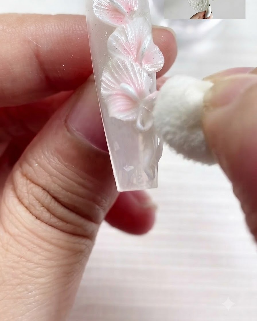 Una pequeña esponja cosmética aplicando suavemente pigmento blanco perla en los bordes texturizados de lirios calla blancos esculpidos y un lazo en una uña.