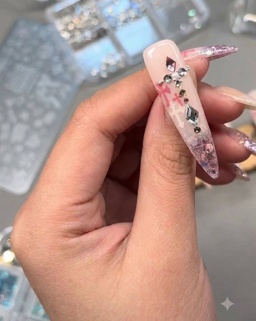 Primer plano de una uña decorada adornada con cristales, una perla y dijes de metal sobre purpurina rosa y arte estampado.