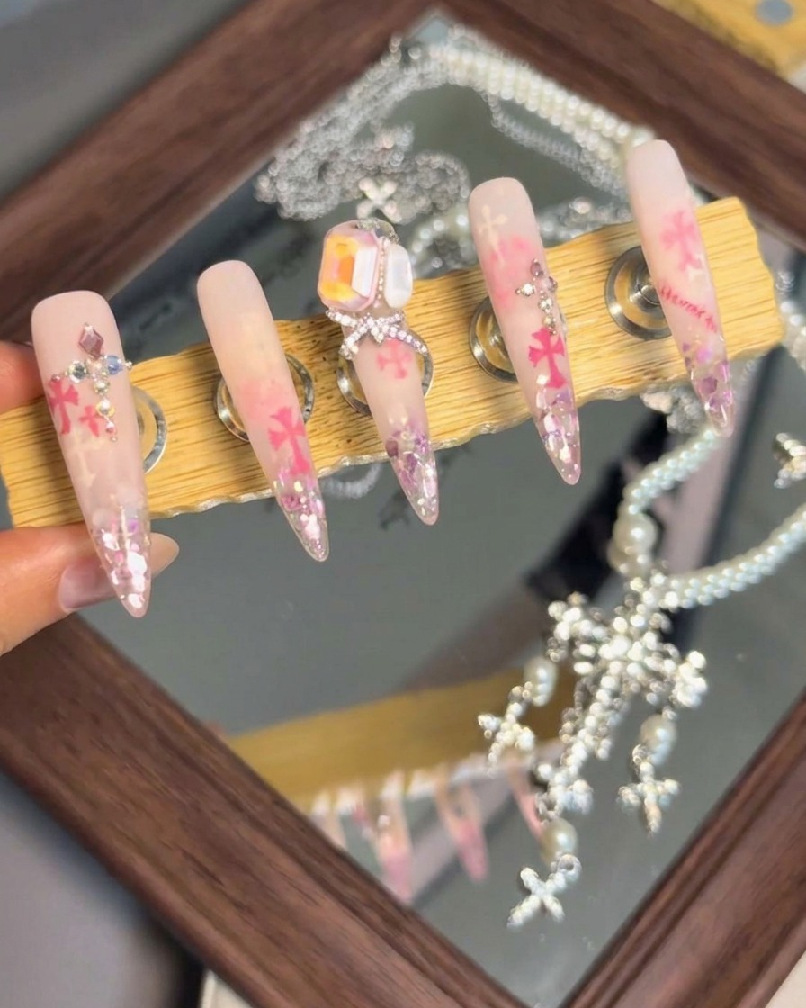 Un soporte de exhibición que muestra cinco uñas postizas terminadas con arreglos de cristales elaborados, lazos y detalles de Chrome Hearts.