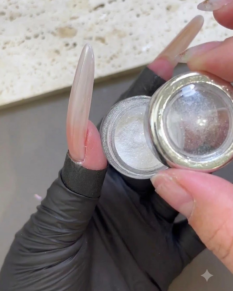 Un dedo con guantes sosteniendo un frasco abierto de polvo iridiscente blanco de luz de luna junto a una uña extendida brillante.