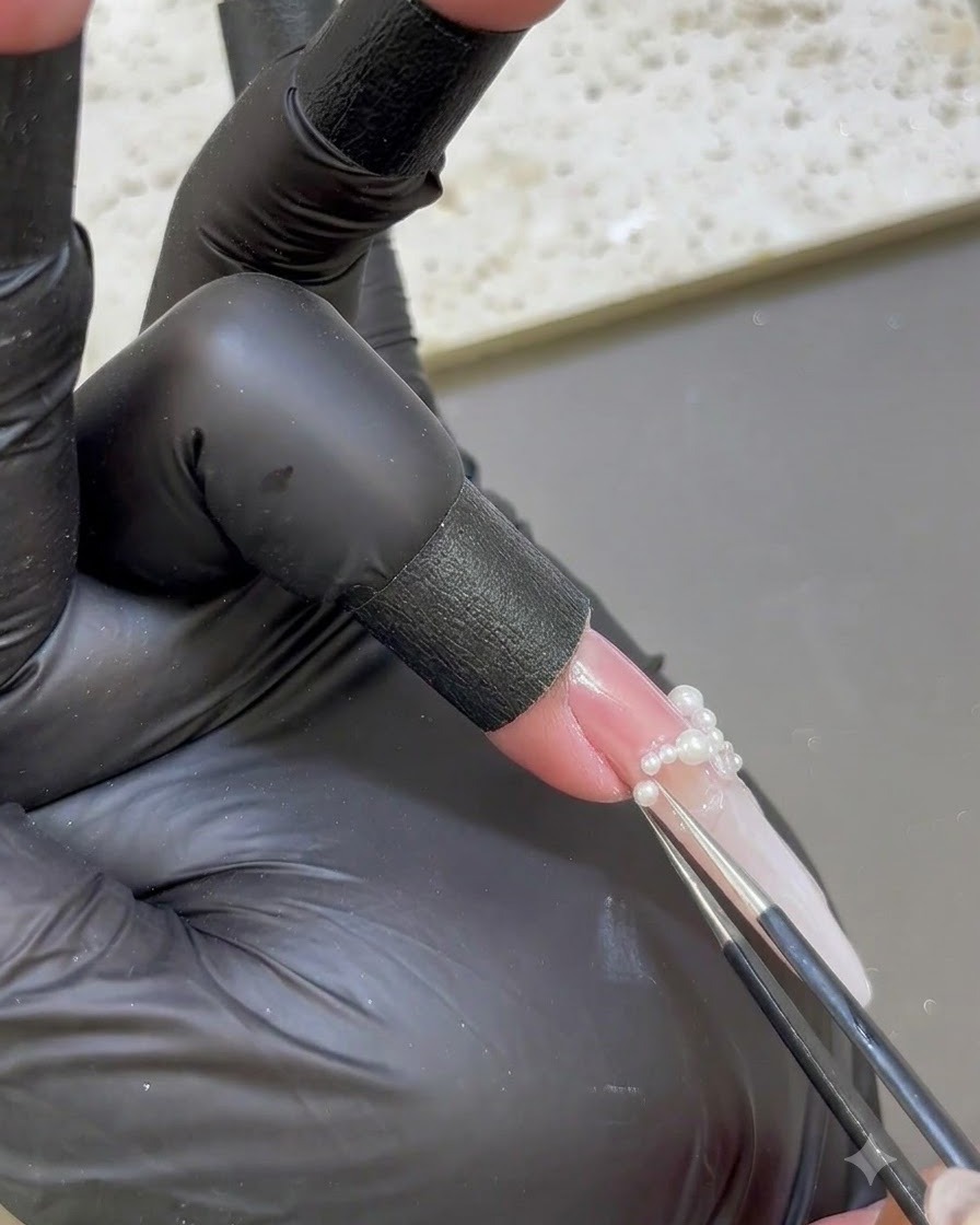 Pinzas de precisión colocando diminutas perlas blancas en la base de una uña extendida.