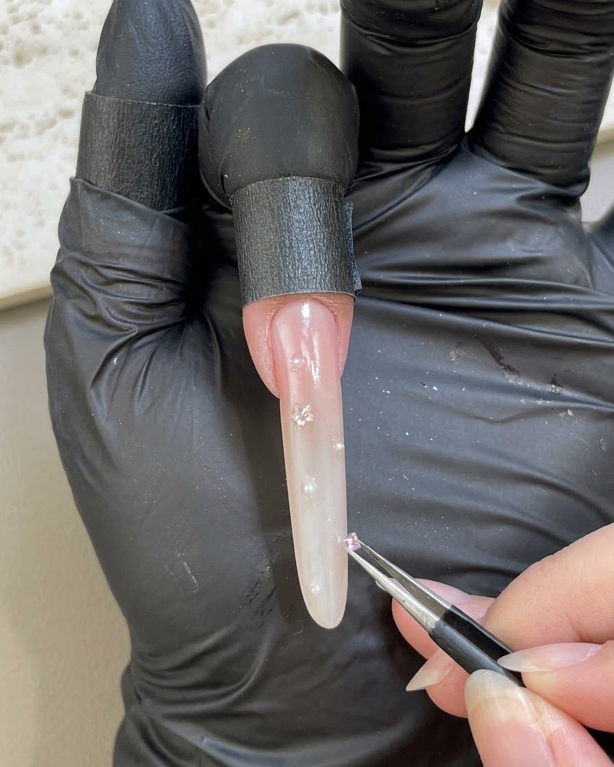 Una mano con guantes usando pinzas para colocar una pequeña piedra de pedrería rosa en una superficie de uña embellecida.
