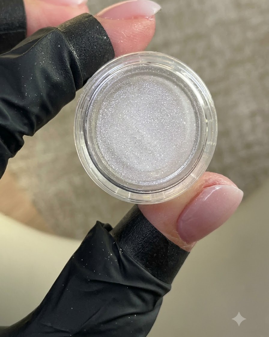 Una mano enguantada sosteniendo un pequeño frasco abierto de polvo blanco brillante con una uña manicurada a la vista.