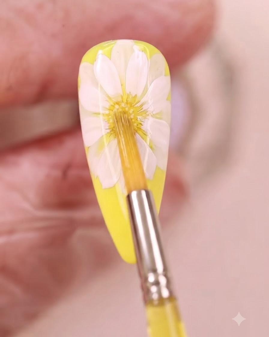 Un pincel fino aplicando líquido de acuarela amarilla en el centro de un diseño de uñas de margarita blanca.