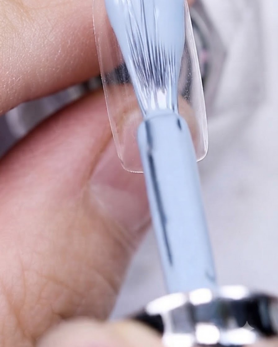 Un pincel aplicando esmalte en gel azul grisáceo sobre una uña natural preparada.