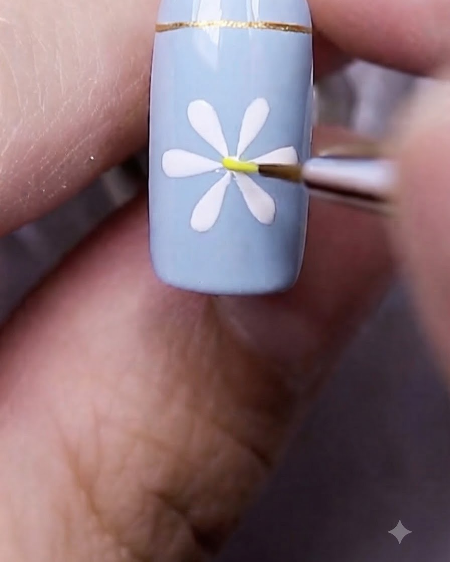 Un pincel fino de nail art colocando un diminuto punto amarillo exactamente en el centro de la margarita blanca.