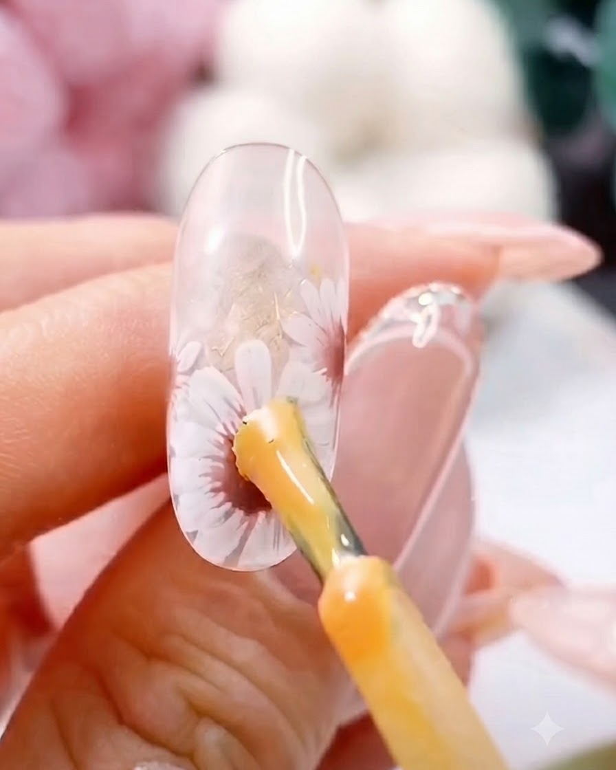 Un pincel aplicando una gota de gel amarillo espeso en el centro de una margarita blanca pintada sobre una uña transparente.