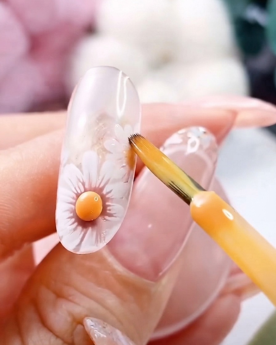 Un pincel fino cerca de un centro de flor amarillo 3D curado sobre una uña de gel transparente.
