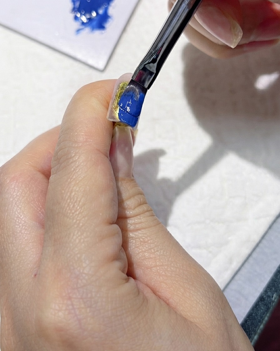 Un pincel aplicando cuidadosamente un esmalte en gel azul oscuro intenso sobre una uña artificial.