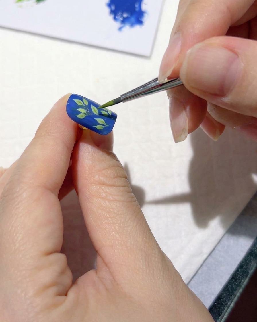 Un pincel fino de nail art agregando trazos de verde claro sobre hojas oscuras en una uña azul.